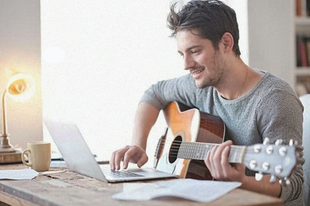 Hombre sonriente tocando una guitarra acústica frente a una laptop sobre un escritorio de madera con una lámpara y una taza