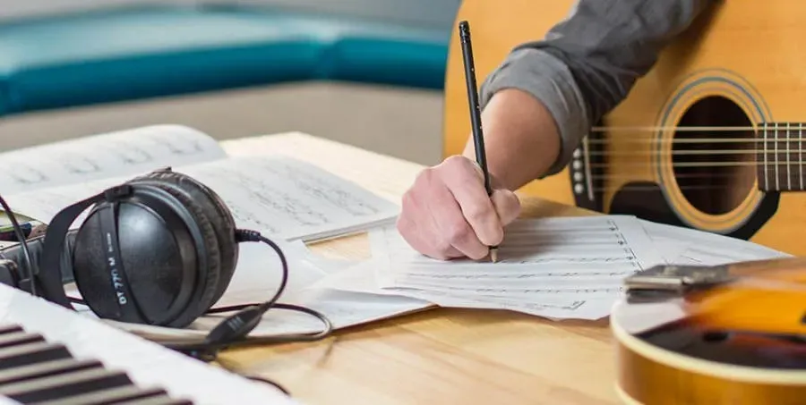 Mano escribiendo una partitura sobre una mesa con audífonos de estudio, un teclado y una guitarra acústica