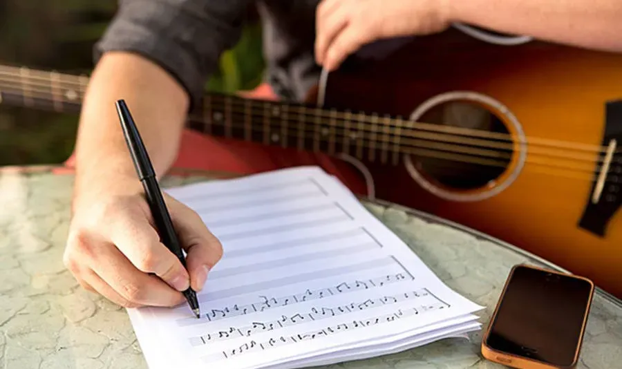 Compositor escribiendo notas en papel pautado con una guitarra acústica y un teléfono sobre la mesa