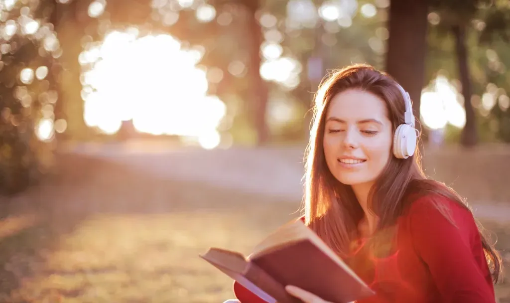 Mujer con audífonos blancos leyendo un libro al aire libre, con los ojos cerrados y la luz del atardecer detrás
