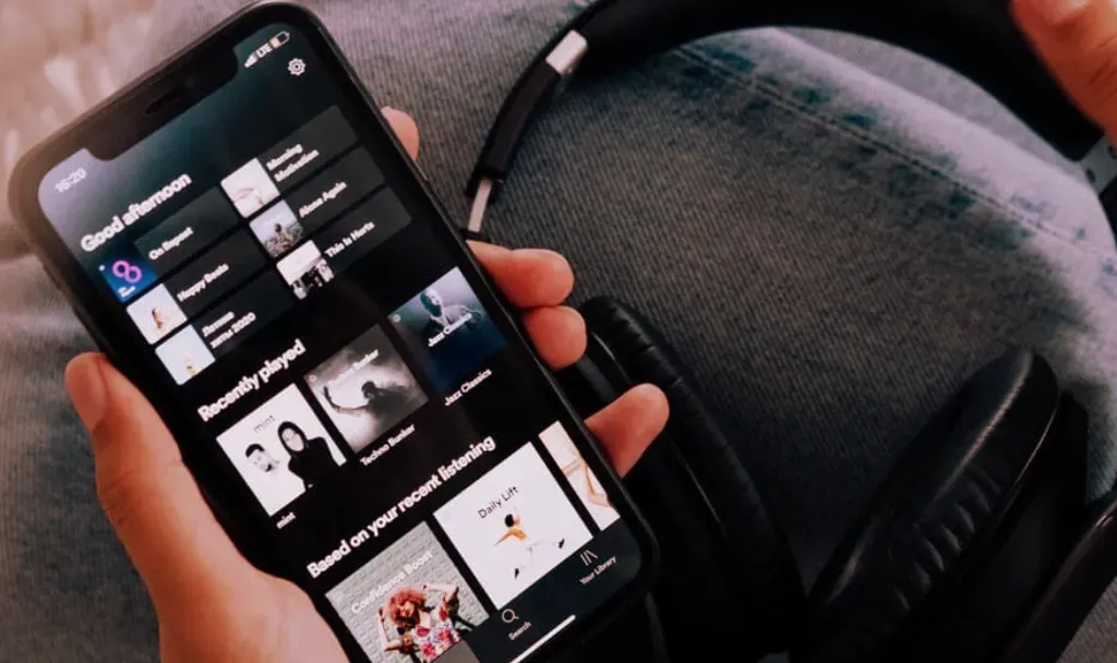 Mano sosteniendo un celular con una aplicación de streaming abierta en la sección de escuchados recientemente, junto a unos audífonos