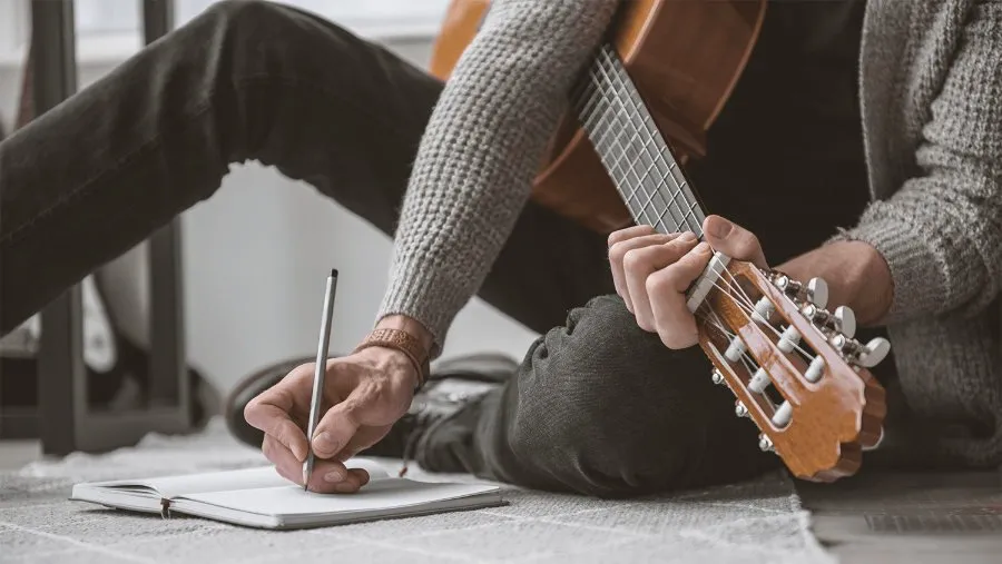 Persona sentada en el suelo escribiendo en una libreta con una guitarra al lado