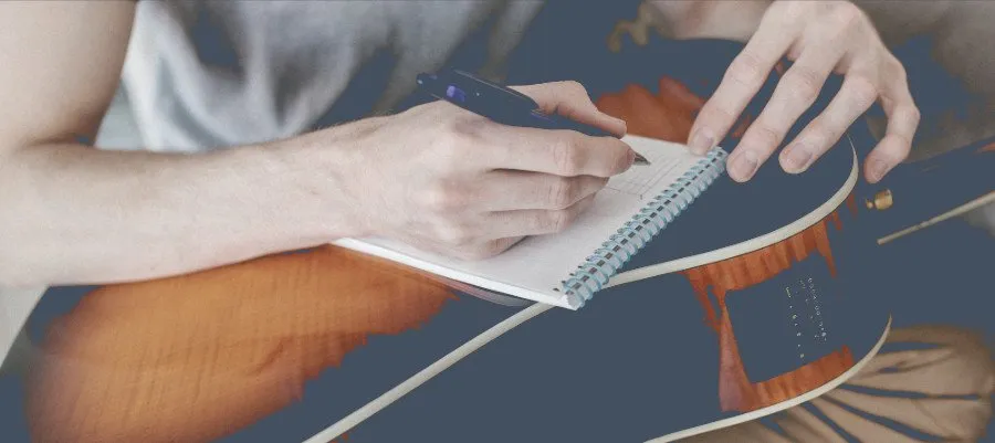 Manos escribiendo la letra de una canción en una libreta apoyada sobre una guitarra