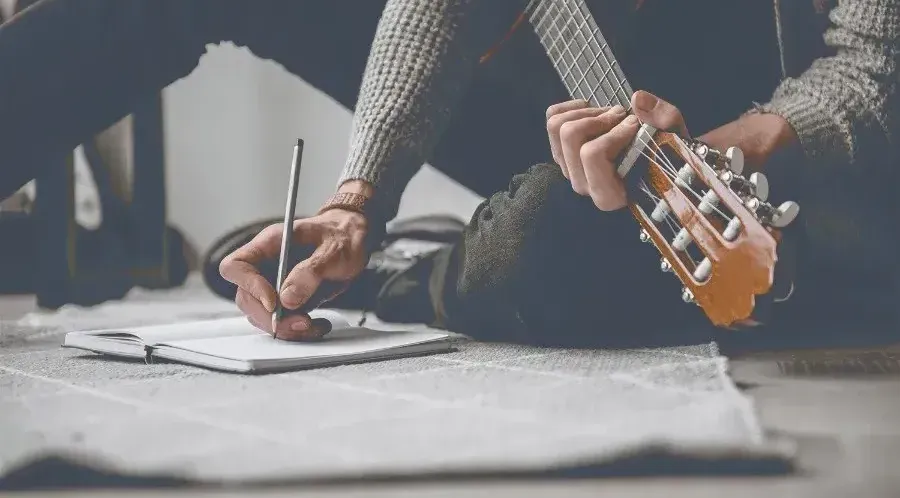 Persona escribiendo la letra de una canción en una libreta junto a una guitarra