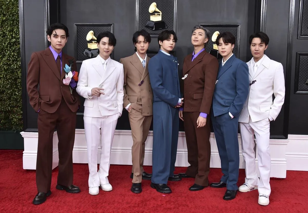 Los siete integrantes de BTS con trajes de colores posando en la alfombra roja de los Grammy, con gramófonos dorados de fondo