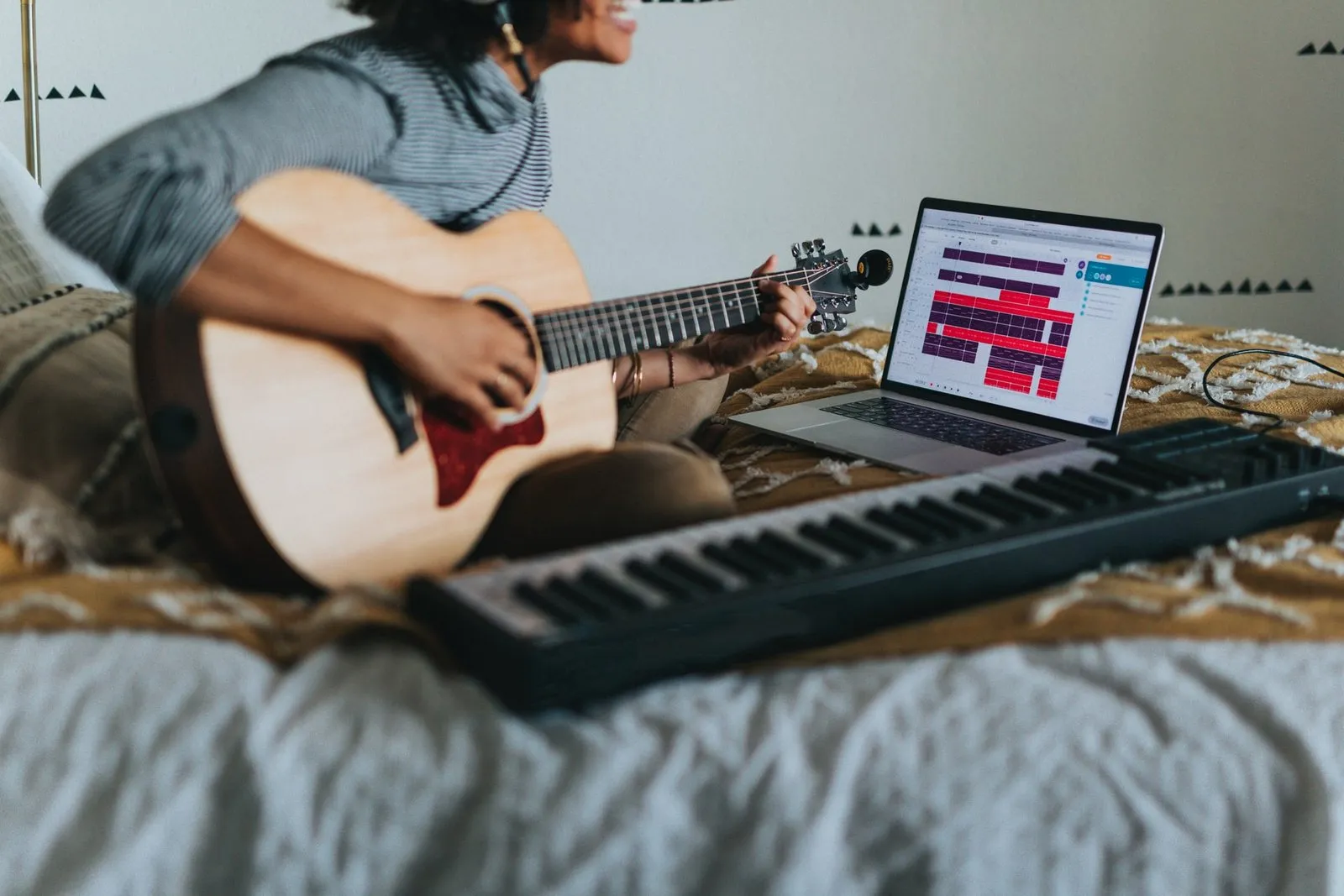 Mujer sonriente toca una guitarra acústica sentada en la cama, junto a un teclado y una laptop con una sesión de grabación abierta