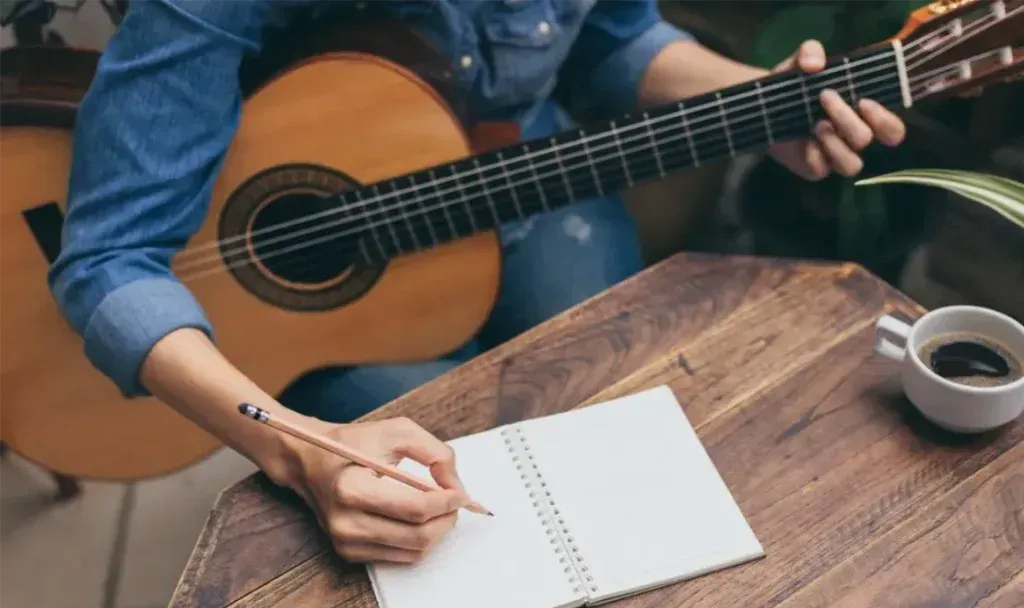 Músico con guitarra criolla escribiendo con lápiz en una libreta sobre una mesa de madera junto a una taza de café
