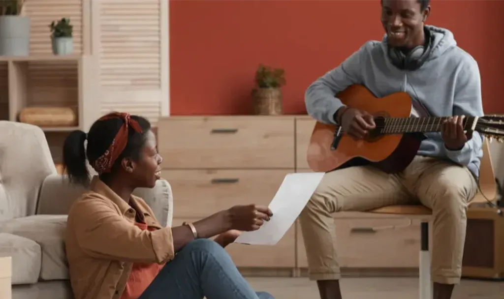 Mujer sentada en el suelo entrega una hoja de papel a un hombre que toca la guitarra acústica en una sala