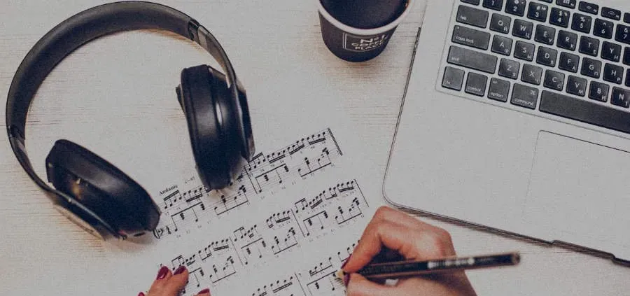 Audífonos, una partitura y una computadora portátil sobre una mesa mientras alguien escribe una canción