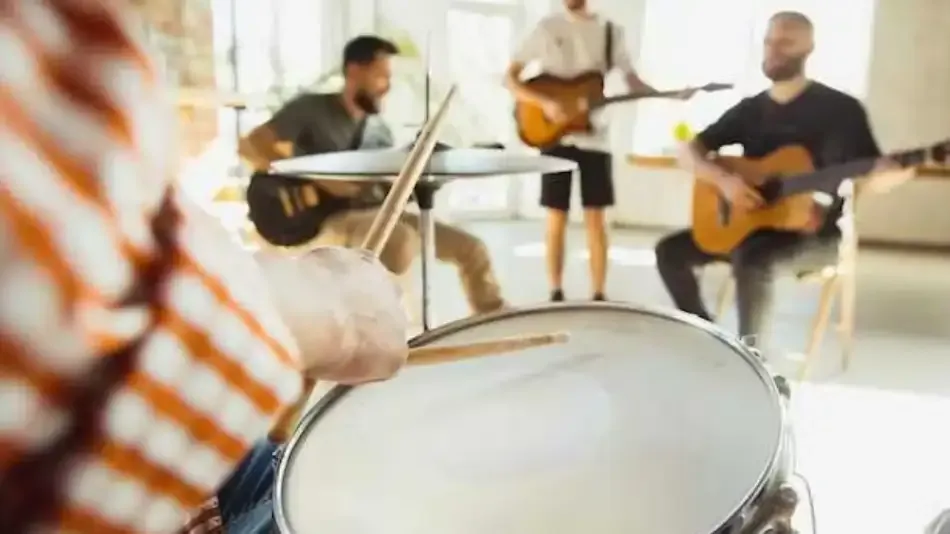 Baterista en primer plano con músicos tocando guitarras eléctrica y acústica al fondo durante un ensayo