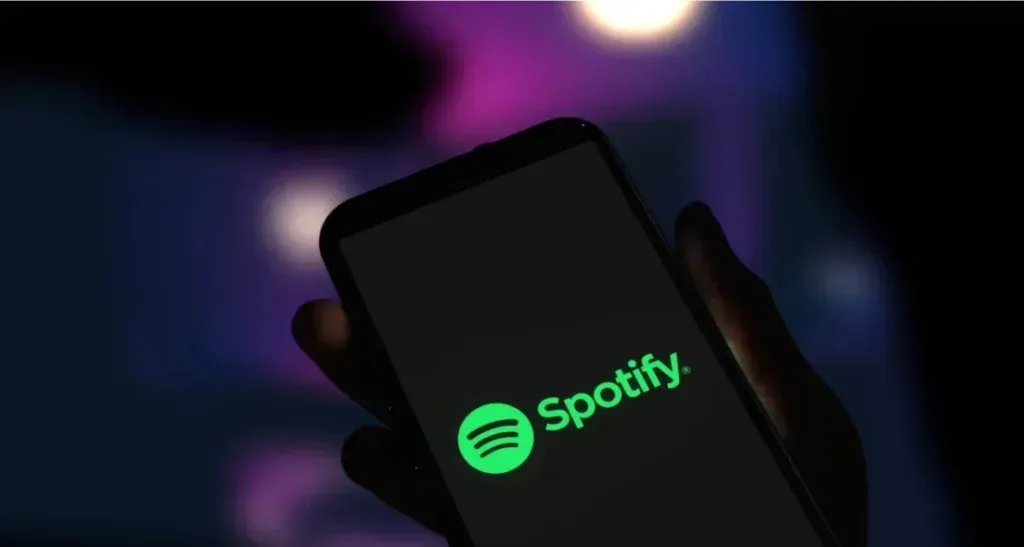 Mano sosteniendo un teléfono con el logotipo de Spotify en la pantalla, sobre un fondo de luces desenfocadas