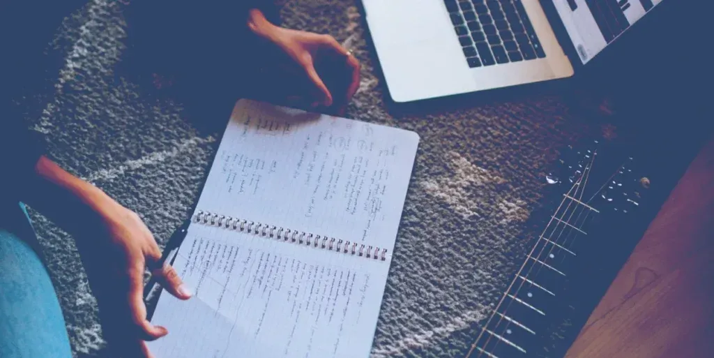 Manos sobre una libreta de espiral llena de letras manuscritas, junto a una laptop y una guitarra eléctrica en el suelo