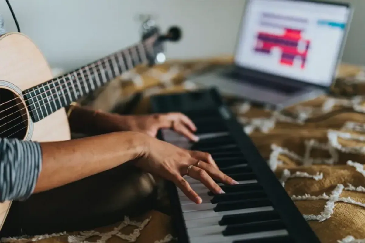 Persona tocando un teclado con una guitarra acústica en el regazo y una laptop con un proyecto musical abierto